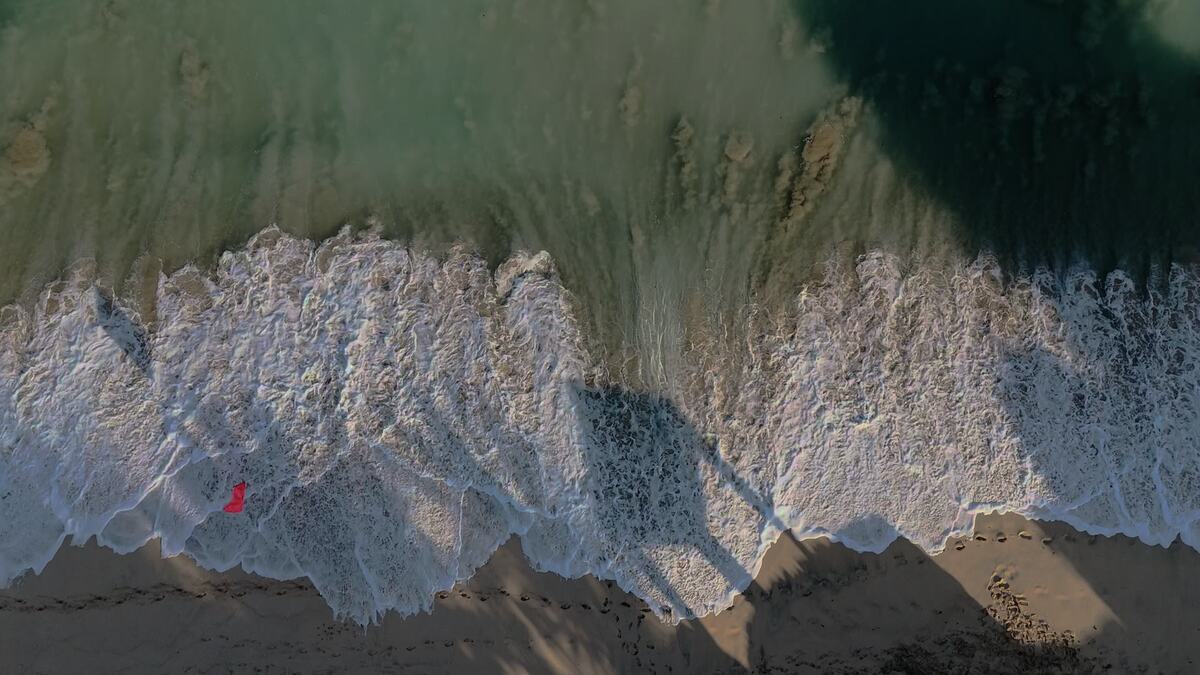 A person walking at the edge of the surf on a Bukit beach, seen from a drone