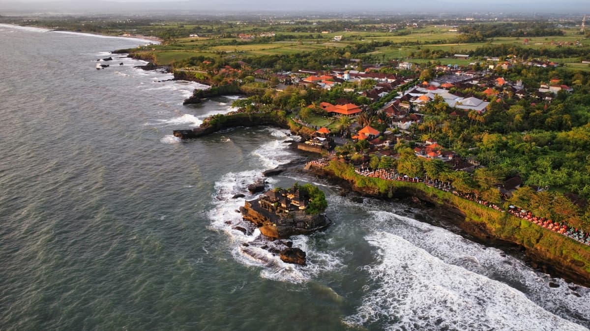 Pura Tanah Lot di atas karang di tepi laut, dengan pesisir Tabanan, sebuah desa dan sawah di belakangnya, difoto dari drone